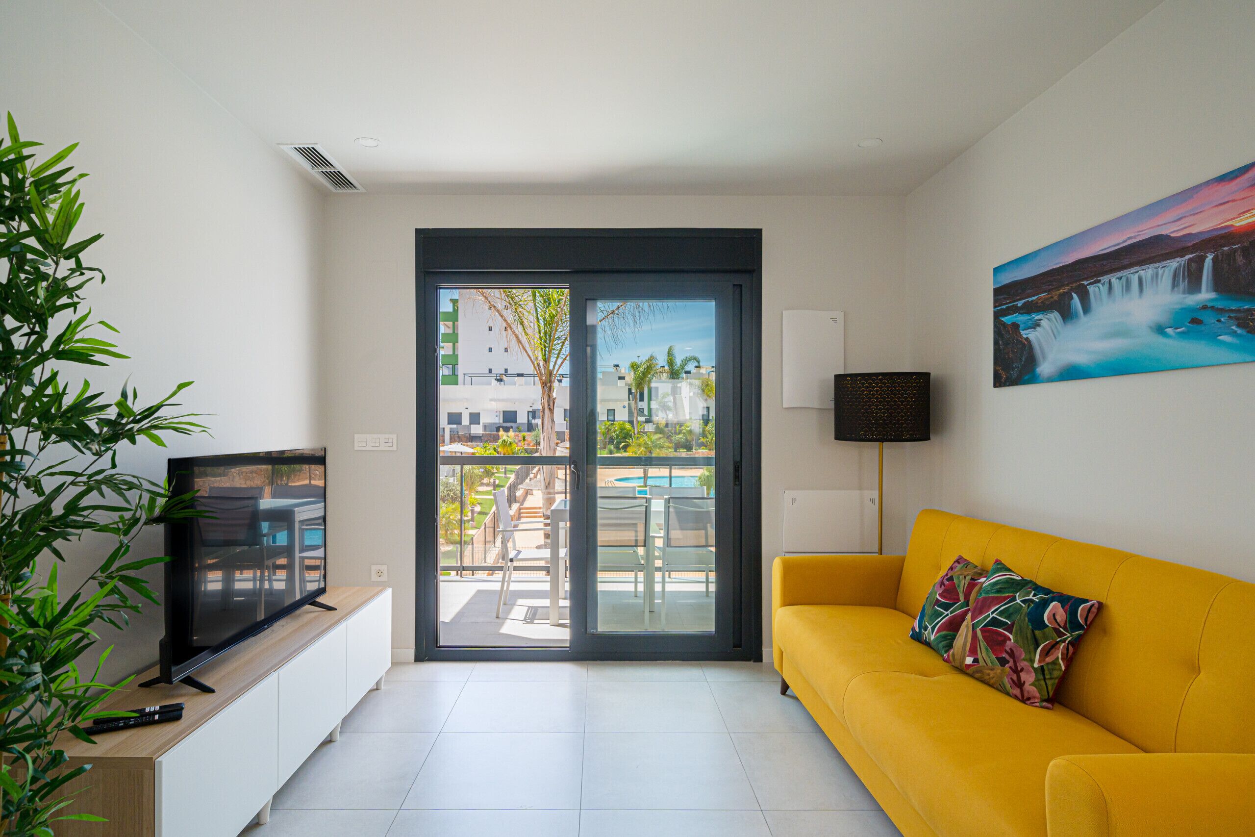 A modern living room with a yellow sofa, a television on a wooden cabinet, and a large window overlooking a pool and buildings.