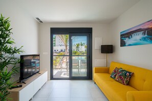 Een felle gele bank, een televisie op een houten kast, een groot raam met uitzicht op een zwembad en gebouwen, en een schilderij van een waterval aan de muur.