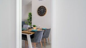 A dining area with a wooden table, two grey upholstered chairs, a round wall clock, and a potted plant. - Apartamento Iseo (Mil Palmeras) (Pilar de la Horadada)