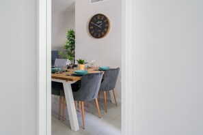 Een eetgedeelte met een houten tafel, twee grijze fauteuils, een ronde wandklok en een potplant.