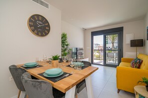Een moderne eetruimte met een houten tafel, grijs gestoffeerde stoelen en een gele bank. Een grote wandklok en een televisie zijn zichtbaar.
