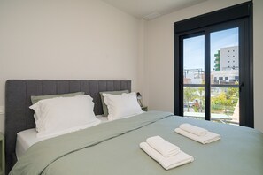 Hotelkamer met een bed, twee kussens, een groene spreideken en uitzicht op gebouwen door een raam.