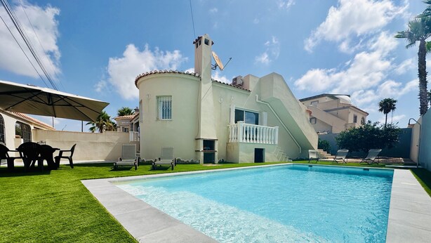 Une piscine avec une terrasse blanche, un espace patio couvert et un bâtiment doté d’un balcon.