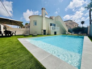 Ein Schwimmbad mit weißer Liegefläche, einer überdachten Terrasse und einem Gebäude mit Balkon.