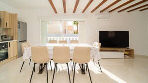 Une cuisine moderne avec des meubles en bois, un four encastré et un réfrigérateur. On y trouve un coin repas avec une table et des chaises, ainsi qu’un grand téléviseur à écran plat posé sur un meuble en bois.
