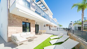 Un espace de vie extérieur moderne, avec un canapé blanc, une table en bois et des fauteuils de jardin blancs, le tout installé sur une pelouse artificielle, à côté d’un bâtiment aux murs en pierre et doté d’un balcon.