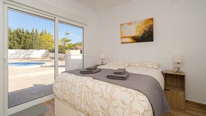 A bedroom with a bed, a nightstand, a painting, and a view of a swimming pool and trees.