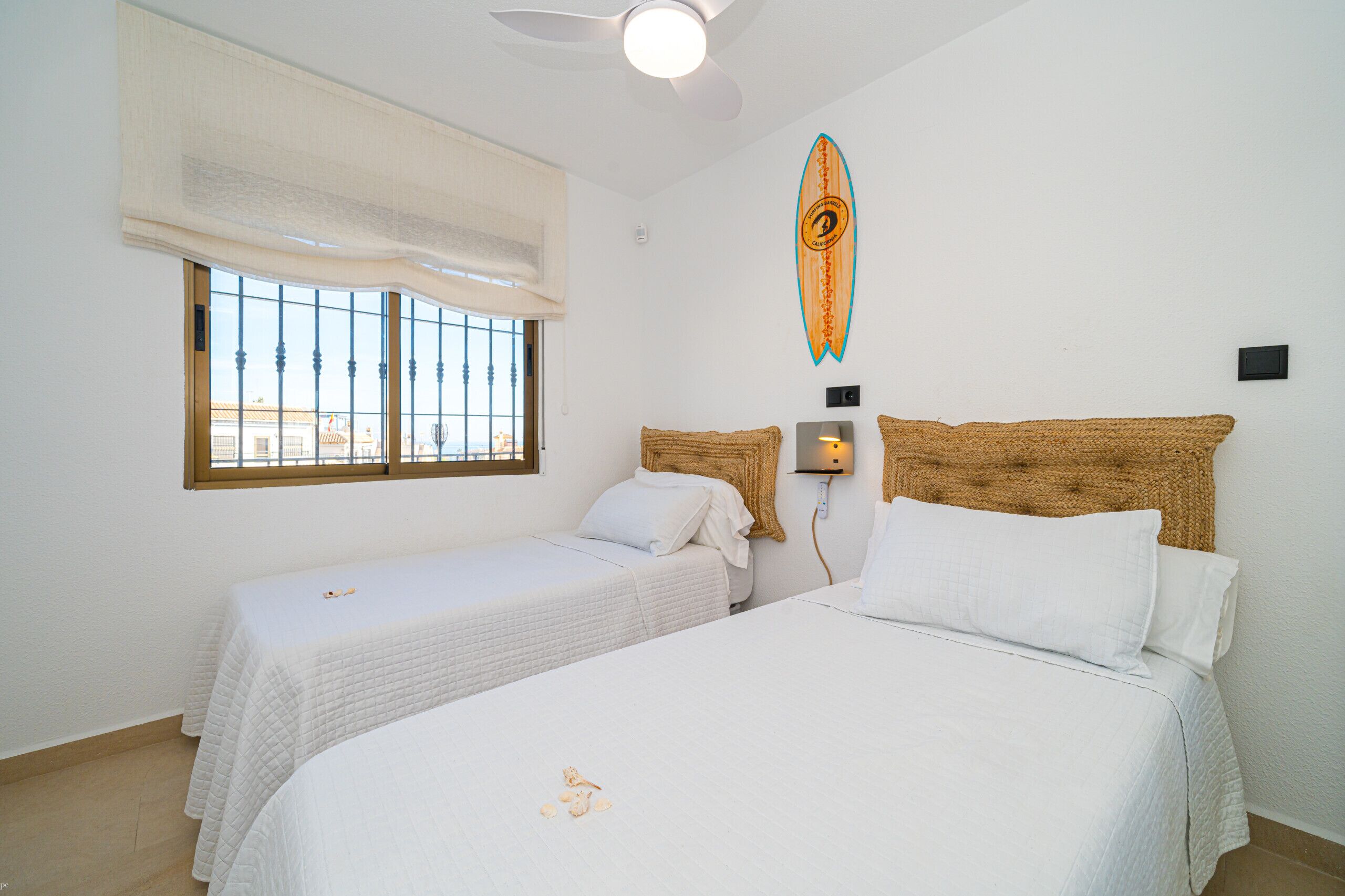 A hotel room with two beds, a ceiling fan, a window with blinds, and a surfboard decoration on the wall.