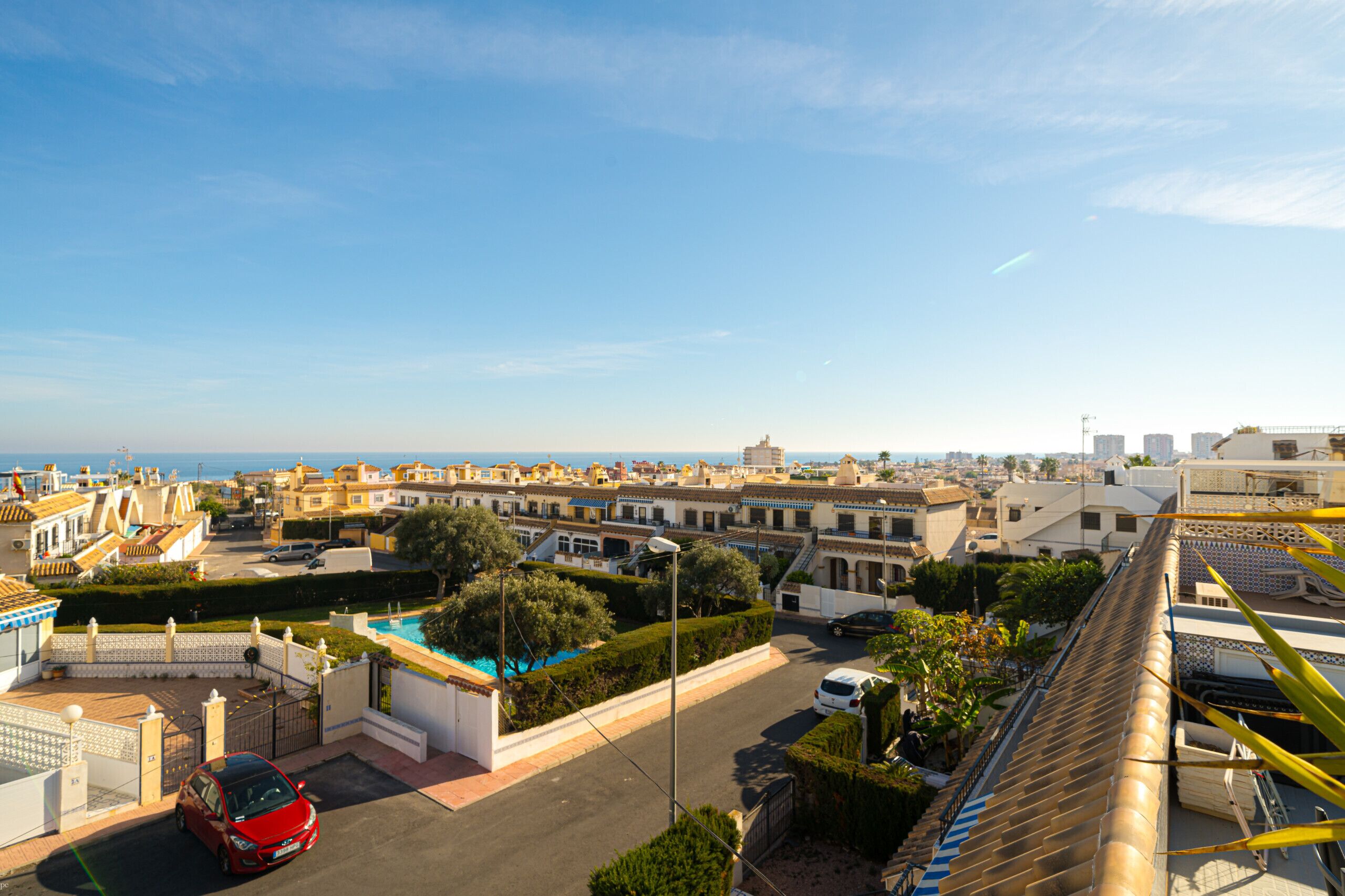 A residential area with a red car parked in a driveway, a swimming pool, and a view of the sea in the background.