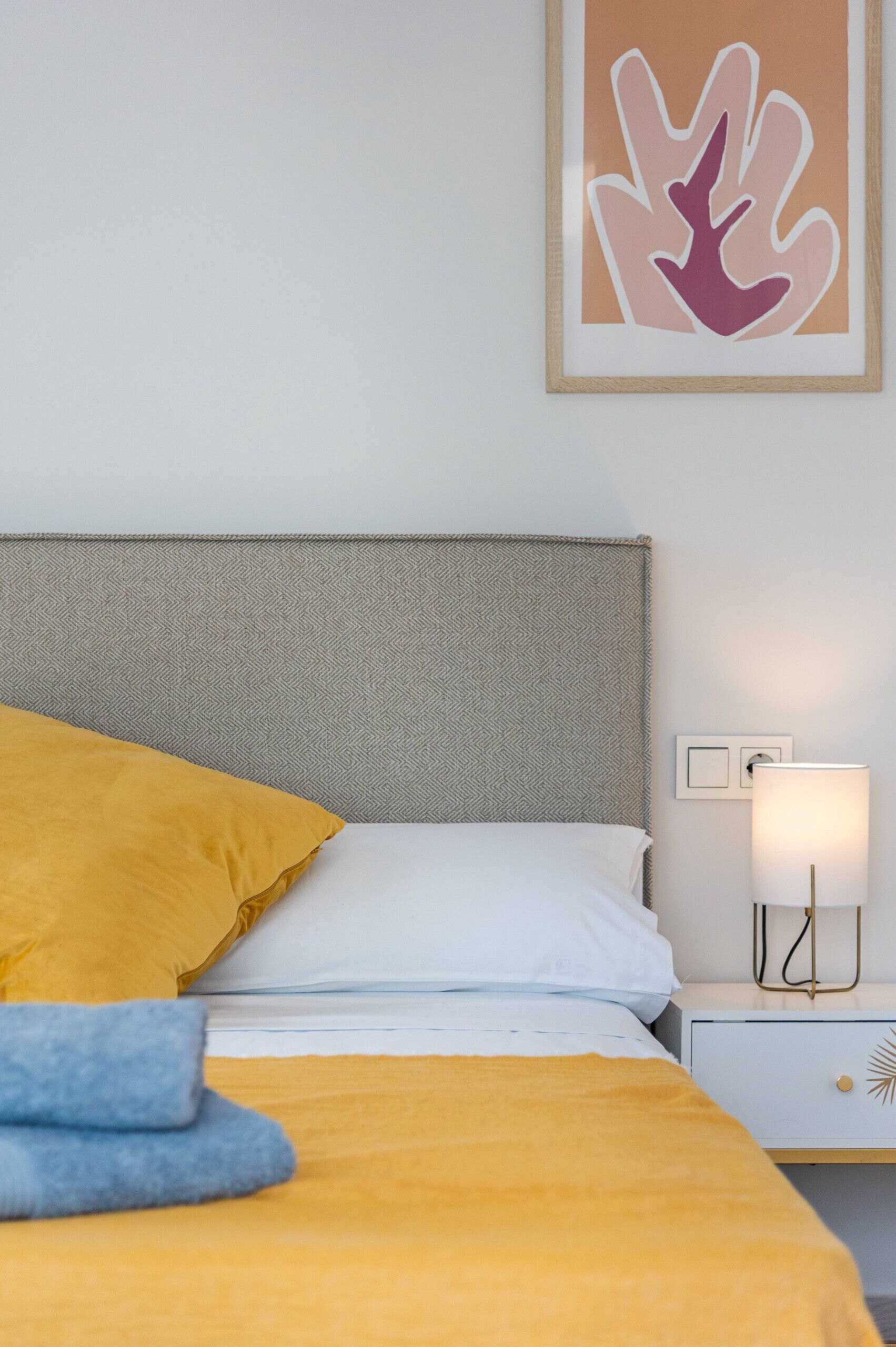 A neatly made bed with a yellow blanket and a single blue towel, a white pillow, and a gray headboard. There is a framed artwork above the bed and a bedside lamp on the nightstand.