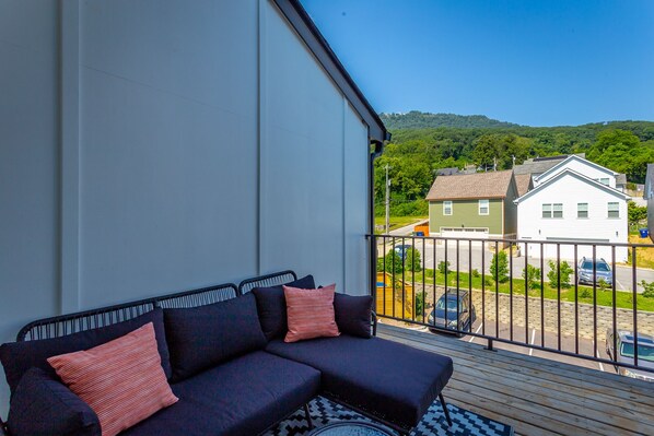 A balcony with a dark-colored sofa and patterned rug, overlooking a residential street with houses and a clear blue sky. - Basecamp - a PMI Scenic City Vacation Rental (Chattanooga)