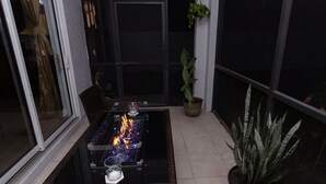 A glass-top table with a fire feature, a potted plant, and a wicker chair. - Boho Gator Lux 2 Ft. Myers (Lehigh Acres)