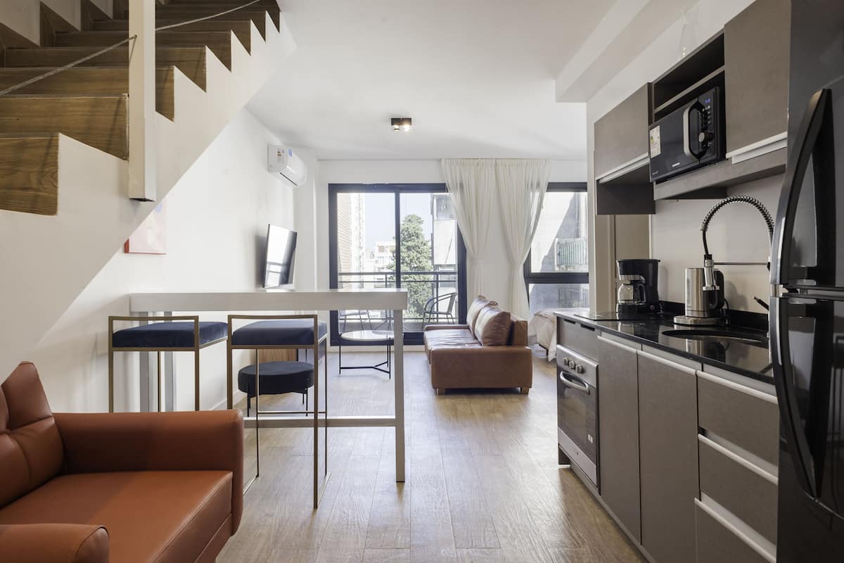 A modern apartment with a kitchen, dining area, and living room. It features a staircase, a sofa, a dining table with chairs, and a large window with curtains.