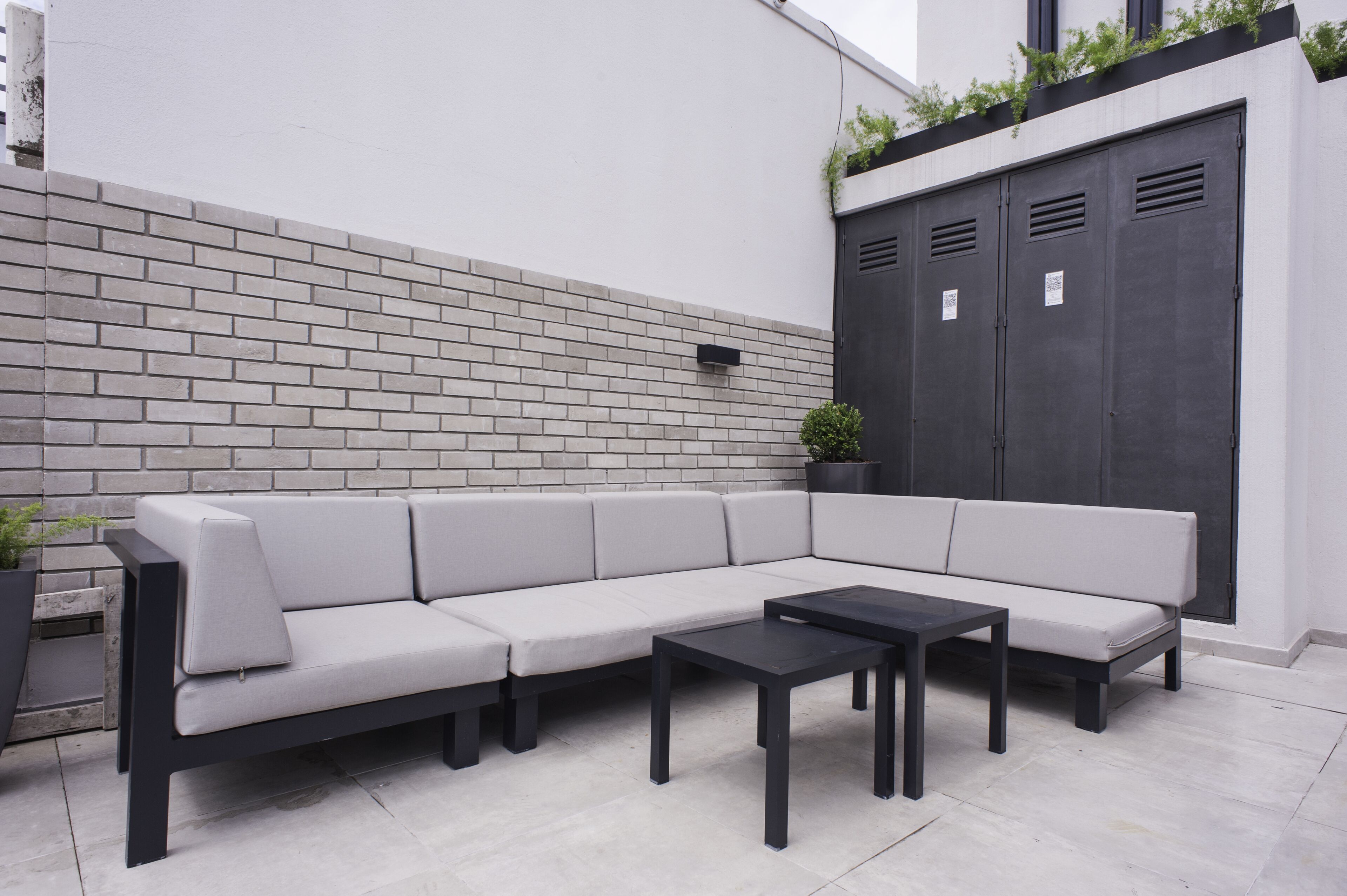 Un espace extérieur moderne, aménagé avec un canapé de couleur claire et deux tables noires, le tout adossé à un mur de briques blanches et à une porte sombre.