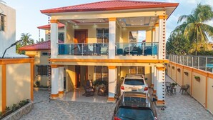 A two-story house with a balcony, a car parked in front, and a pool in the background.