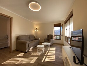 Ein modernes Wohnzimmer mit einer Couch, zwei Beistelltischen, einem Fernseher und einem großen Fenster mit Ausblick.