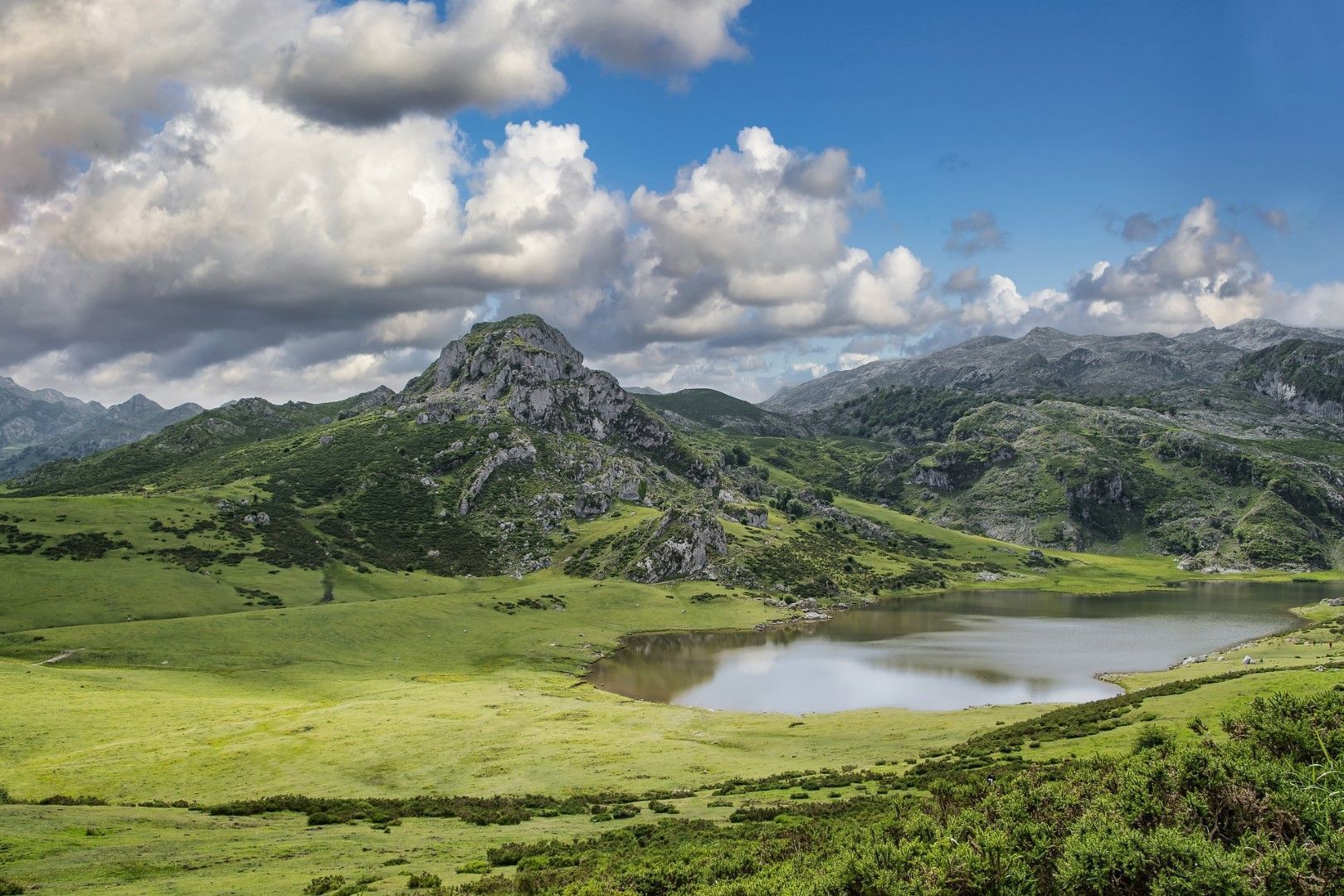 A mountainous landscape with a prominent rocky peak, a lake, and rolling green hills.