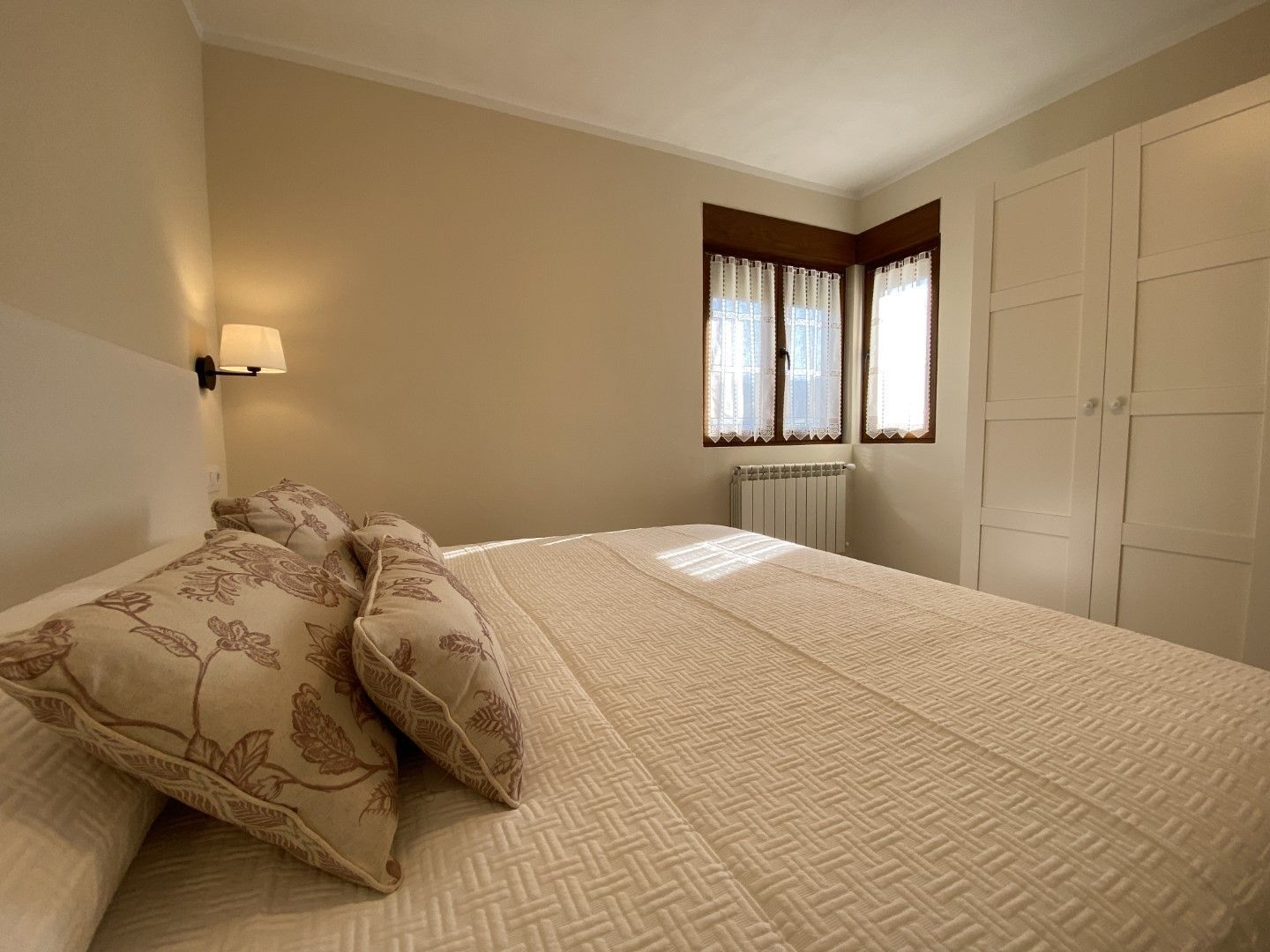 Ein Schlafzimmer mit einem Bett, Kissen mit Blumenmuster, einer Wandleuchte, einem Heizkörper und einem Fenster mit Vorhängen.