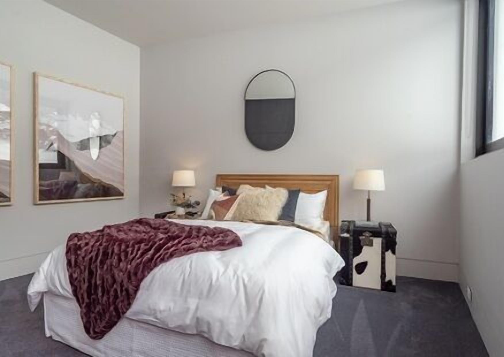 Una moderna camera d'albergo con un letto grande, due comodini con lampade, un grande specchio sopra il letto e due opere d'arte incorniciate a parete.