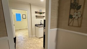 A hallway with a kitchen area in the background, a door leading to another room, and a wall with a floral decoration.