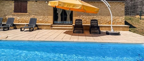 En swimmingpool med ett uteområde, inklusive en möblerad, delvis takförsedd sittplats med stolar och ett parasoll.