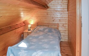 A small, narrow room with wooden walls and a sloped ceiling. There is a bed with a floral bedspread and a nightstand with a lamp.