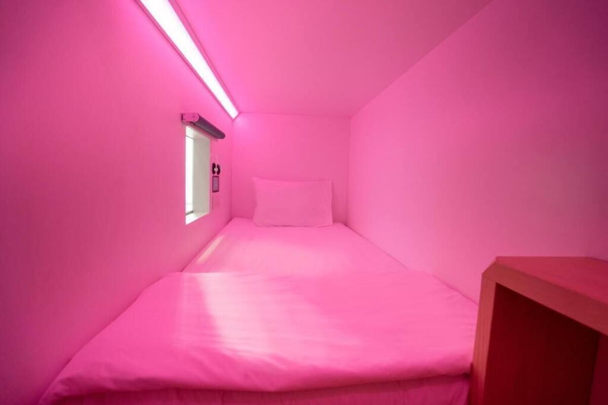 A small, pink-themed room with a single bed, a window, and a wooden nightstand.