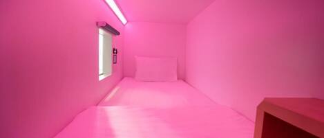 A small, pink-themed room with a single bed, a window, and a wooden nightstand.