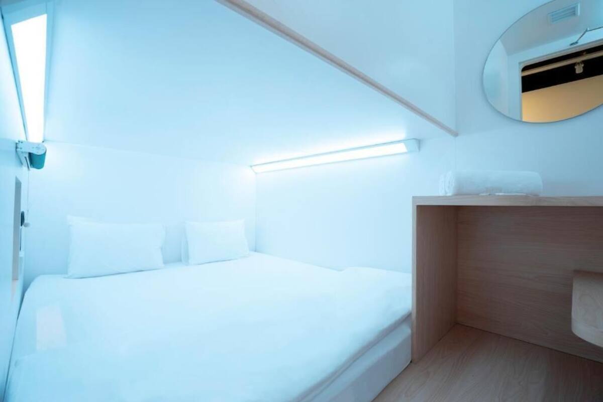 A modern, minimalist bedroom with a bed, a wooden nightstand, and a wall-mounted mirror.