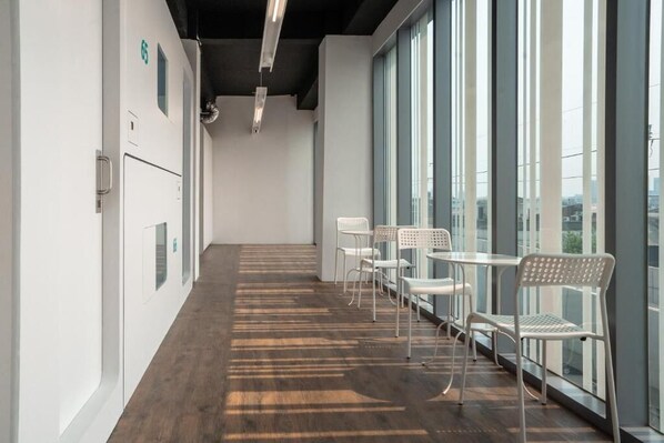 A modern hallway with a wooden floor, white walls, and large windows. There are white chairs and tables arranged along the windows.