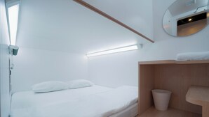 A minimalist bedroom with a bed, a bedside trash bin, and a small wooden shelf. - Bobopod Kebayoran Baru Jakarta (Jakarta)