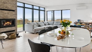 A modern living room with a fireplace, a large sofa, a dining table with chairs, and large windows offering a view of the outdoors.