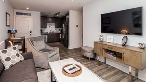 A modern living room with a sofa, coffee table, and TV. There is a kitchen in the background with a fridge and microwave. - Hotty Toddy Condo at 514 by Apex Short Term Stays (Oxford)