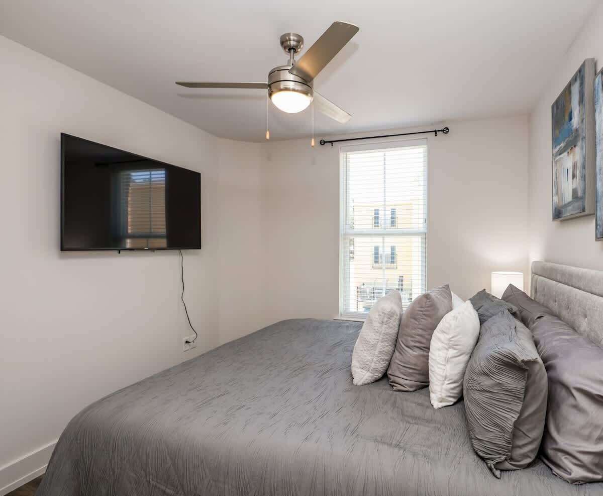 A bedroom with a bed, a ceiling fan, a TV mounted on the wall, and a window with blinds.