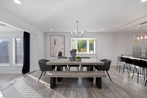 A modern dining area with a rectangular table, black chairs, a bench, a chandelier, and a large window with blinds.