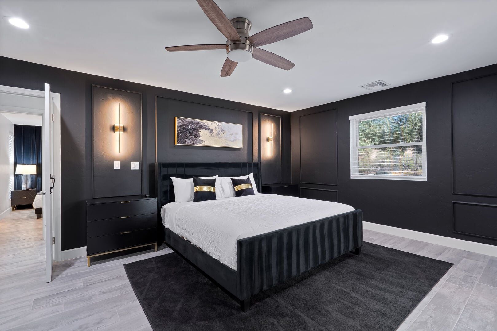 A modern bedroom with a large bed, dark walls, a ceiling fan, and a window with a view.