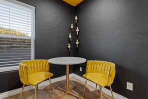 Ein Zimmer mit zwei gelben Polstersesseln um einem runden Tisch, einer schwarzen Wand und einem Fenster mit Jalousien.