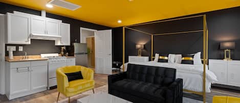 A modern hotel room with a four-poster bed, a separate dining area with a stove, and a yellow armchair.