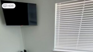 Una sala de estar con un sofá color beige, una mesa redonda de centro, un televisor montado en la pared y una ventana con persianas.