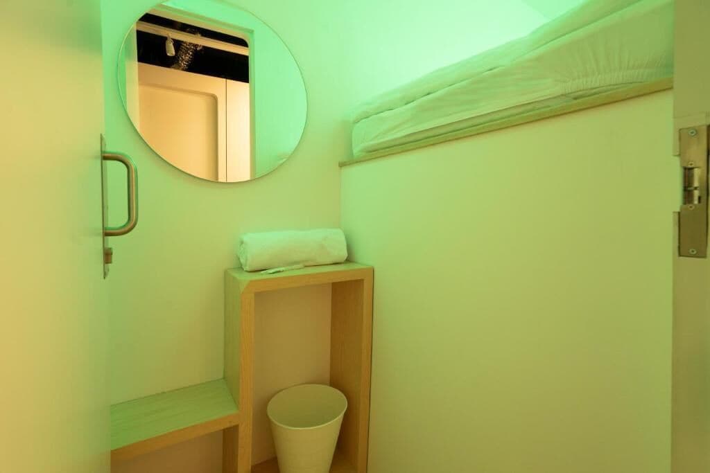 Une petite pièce compacte, équipée d’un lit superposé, d’un miroir rond, d’une étagère en bois et d’une poubelle.