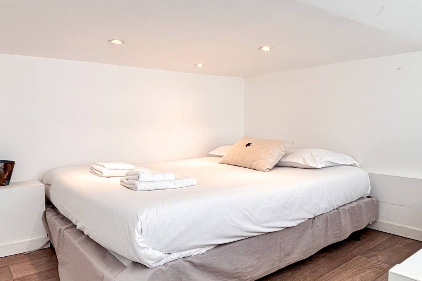 A neatly made bed with white linens and a single pillow, a bedside table with a book, and a wooden floor.