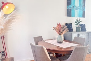 Un coin repas avec une table ronde, six chaises, un vase contenant des fleurs séchées, une lampe et un tableau accroché au mur.