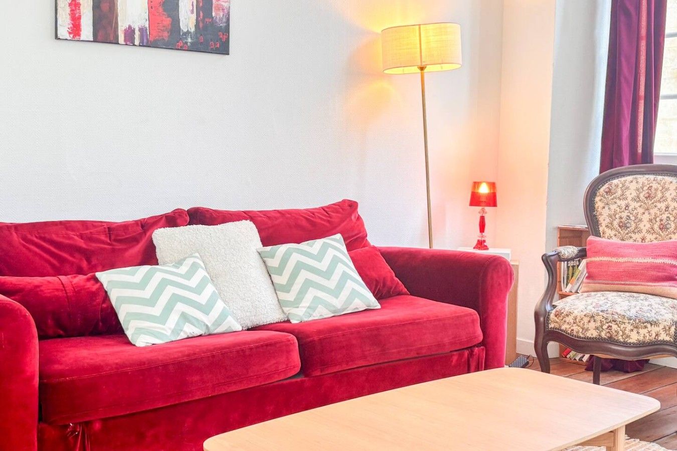 A red sofa with patterned pillows, a wooden coffee table, a floor lamp, and a patterned armchair.
