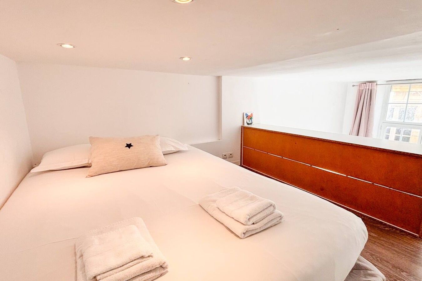 A hotel room with a bed, a headboard, a window with curtains, and a bedside table.