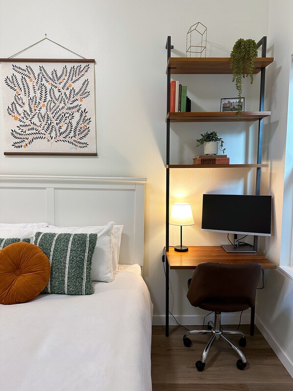 A neatly arranged bedroom with a bed, bedside table, lamp, and a desk with a computer monitor.