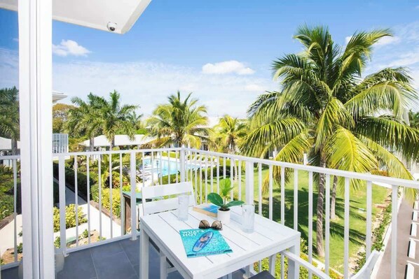A balcony with a white table, a chair, and a view of a pool and palm trees.