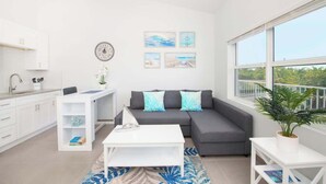 Een moderne woonkamer met een grijze bank, een witte salontafel, en een eethoek met een witte tafel en stoelen. Er hangen afbeeldingen in lijsten aan de muur en een groot raam biedt uitzicht op de omgeving.