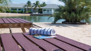 Mesa de picnic de madera con una toalla enrollada y una revista, con vista a una playa con palmeras y una casa al fondo.