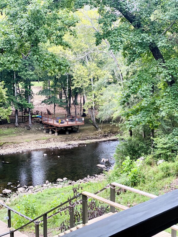 A wooden deck overlooking a river with a wooden bridge and picnic tables.