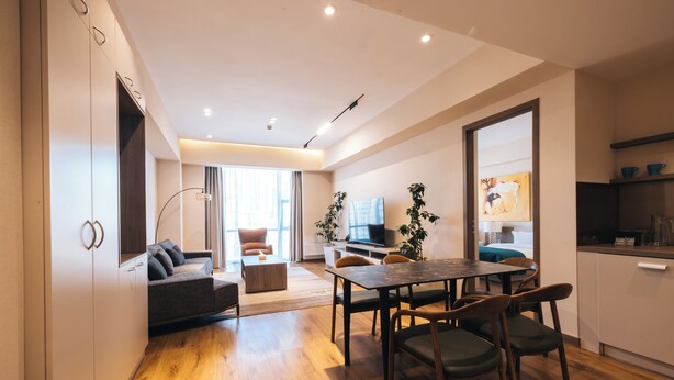 A modern living room with a sofa, dining table, and chairs, a kitchen area with cabinets and a countertop, and a large window providing natural light. - May Seven Hotel and Suites (Ulaanbaatar)